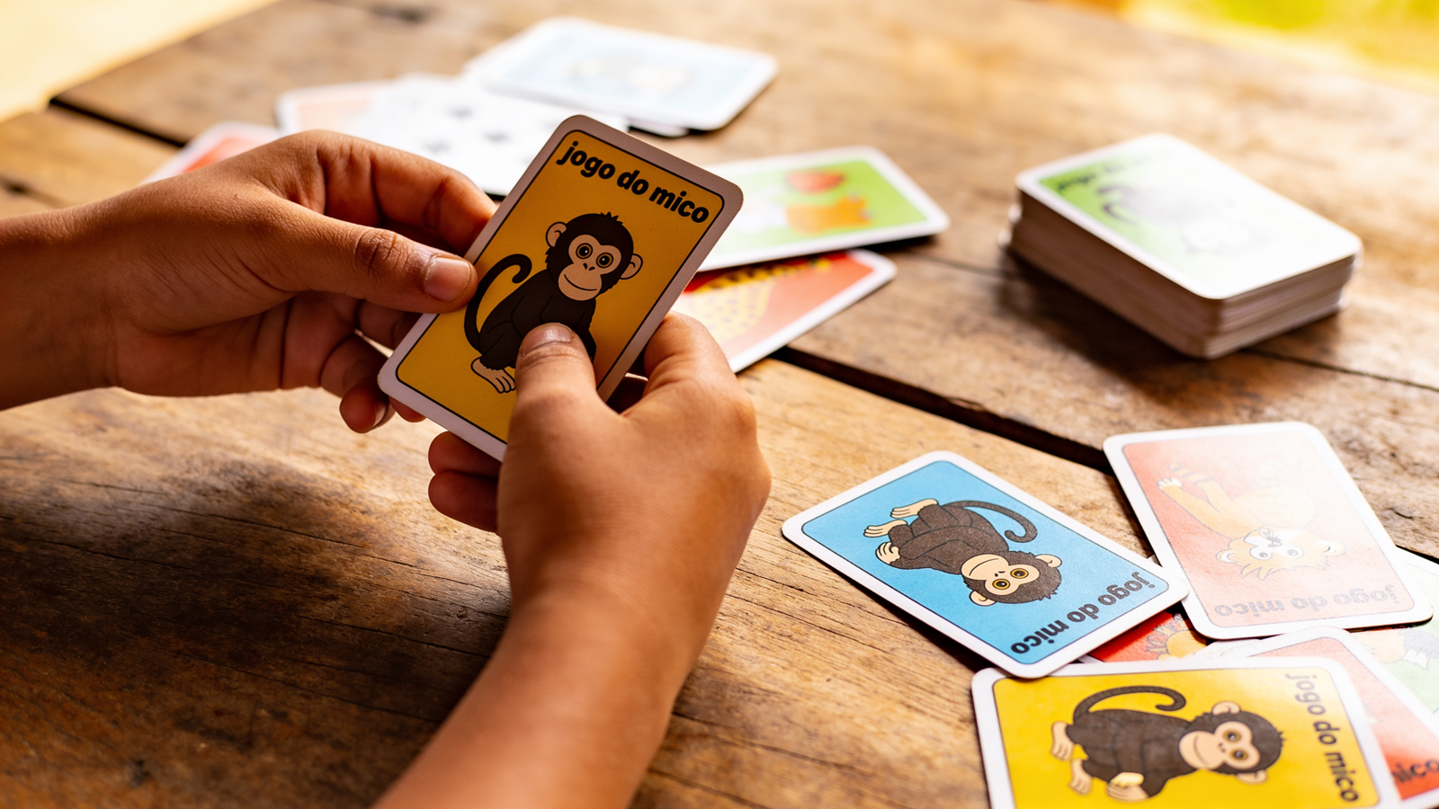 Mãos infantis segurando cartas coloridas do jogo do mico sobre mesa de madeira com baralho espalhado ao redor